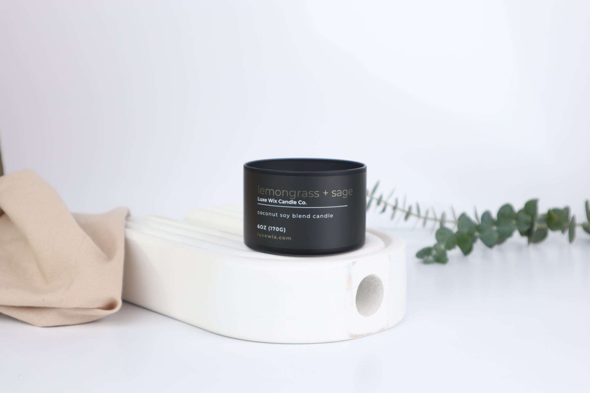 Black candle with a label on a white surface with a beige cloth and green leaves.