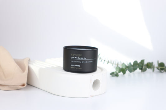 Black candle jar with white label on a white surface with a beige cloth and green leaves.