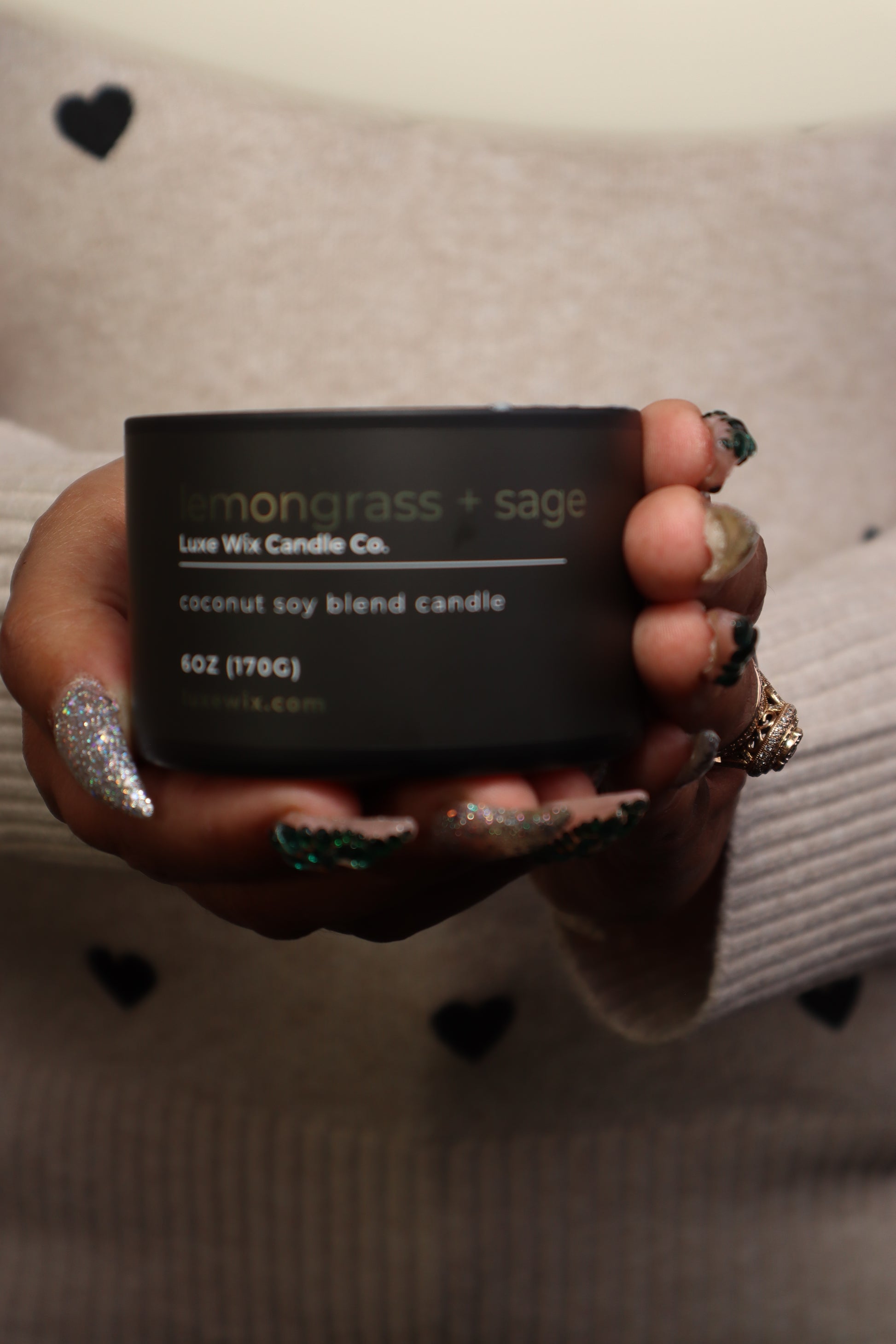 Hand holding a black candle box labeled 'lemongrass + sage' against a neutral background.
